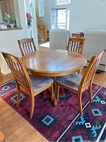 Oak round dining table with four matching chairs arranged around it on a red patterned rug. Table surface looks used with visible marks.