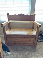 Front-facing view of pine bench showing scalloped backrest, armrests, flat seat with subtle wood grain and knots, and storage panel in front.