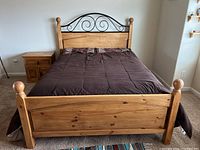 Front view of pine queen bed frame with black iron scrollwork headboard and matching nightstand beside it.