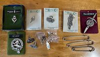 Various Scottish-themed pins, including boxed pin, pins on cards, clan badges, cufflinks, and kilt pins on a wooden surface.