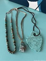 Photo showing all three necklaces laid flat on a blue surface: a turquoise bead and silver bead necklace, a multi-strand silver chain necklace with a stone pendant, and a turquoise shaped pendant on a brown cord.
