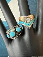 Image showing two rings on black display hand, one with three round turquoise stones, the other a V-shaped ring with turquoise inlay and a clear stone with gold-tone leaf detailing.