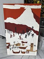 Framed textile fabric art showing winter village scene with ski figures, trees, houses, and mountain