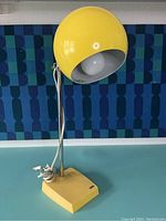 Front angled view of yellow eyeball lamp on blue table against patterned textile with visible plug and cord wrapped at the base.