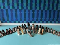 Wide angled photo showing about 30 miniature shoes arranged in a curved row on a blue surface against a blue patterned background. Shows different styles including boots, heels, and dress shoes mostly metallic or painted metal/plastic.