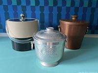Three mid-century style metal ice buckets arranged on a blue surface with geometric blue background.