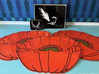 Five bright orange vintage plastic poppy placemats spread out with a black and white Jason Weiss Australia box set placemat and individual coaster behind them on blue surface.