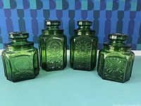 Four green glass Wheaton canisters with lids, arranged side by side showing front embossed sunburst floral motifs and geometric paneling.