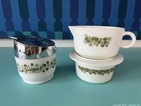 Three vintage Pyrex Spring Blossom pieces: two covered sugar bowls with stainless steel lids, and one gravy dish with matching lid and saucer. White opaque glass with green floral pattern.