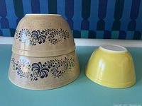 Stacked two tan Homestead Pyrex mixing bowls with dark blue floral pattern alongside a small solid yellow Pyrex mixing bowl on a teal surface and blue patterned backdrop.