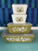 Four stacked Pyrex Spring Blossom refrigerator dishes with glass lids showing white and green floral patterns, on a blue table with a geometric blue background.