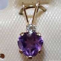 Close-up front view of a yellow gold pendant featuring a heart-shaped purple amethyst and a small round clear stone above it, secured by prongs with a bail for chain attachment.