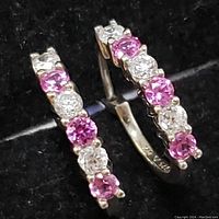 Close-up of pair of sterling silver earrings with alternating created pink sapphire and clear stones, showing prong settings and hinge closure.