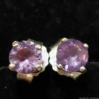 Close-up of the pair of sterling silver amethyst stud earrings laid on a black background, showing details of the purple faceted stones and silver prongs.