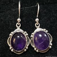 Front-facing view of pair of earrings showing oval cabochon amethyst stones with silver decorative bezel and hooks.