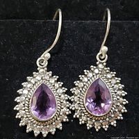 Pair of sterling silver amethyst earrings with pear-shaped stones and detailed silver beadwork surrounded setting, photographed against black background.