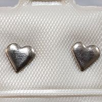 Pair of sterling silver heart shaped stud earrings in a white textured earring holder showing smooth, slightly rounded polished surface with minor scratches.