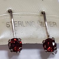Close-up front view of one earring with round faceted garnet stone in a four-prong sterling silver setting, with 'STERLING SILVER' mark visible on the holder.