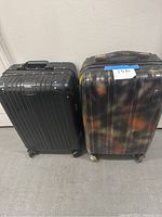 Two carry-on spinner suitcases side by side on floor: black Rimowa on left with broken latch, and a multicolor spinner on right with colorful streaks