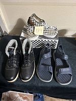 Three pairs of men's shoes displayed on black fabric with some packaging box shown. Pairs include black lined skate-style shoes, black sandals with velcro straps, and black-and-white checkered slip-ons.