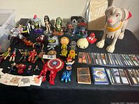 Overview of Magic the Gathering cards, Pokemon cards, Konami cards, multiple figurines, a large Superman dog figure, and some cat-themed temporary tattoos arranged on a black surface.