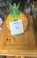 Top view of ceramic pineapple teapot on wooden stand showing teapot lid details and stand surface with inscription.