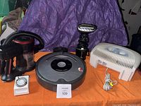 Wide view of all five items on orange cloth with purple backdrop: iRobot Roomba robot vacuum, Homedics steamer, Honeywell fan, Moosoo cordless vacuum, Intermatic timer.