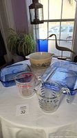 Photo showing the assorted Pyrex items including blue glass baking dishes, vintage white bowl with blue floral design, and two glass measuring cups on a white table near window.