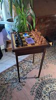 Full view of antique wooden chess table with built-in chessboard and two drawers, with original chess pieces set up and instruction booklet resting on board.