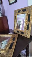 Two matching mirrors with black and gold ornate wood frames on a dark wooden surface, showing their bevelled edges and intricate design.