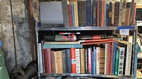 Two metal shelves filled with over 50 books in an indoor space with stone walls and visible pipes.