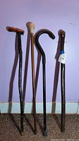 Four assorted vintage canes with varied handles and shaft styles photographed standing upright against a purple wall background on a carpeted floor.