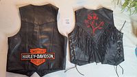 Two black leather women's vests side by side on table. Left vest is Harley Davidson with orange and white patch on back. Right vest is Red Rose with red rose graphic, fringe details, and lace-up sides.