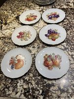 Six porcelain fruit plates with various fruit design and gold trim displayed on granite.