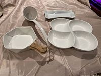 Various white ceramic serving pieces including ladle, four-section dish, rectangular dish, and hexagonal dish with a wooden handle on a fabric surface.