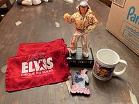 Full display of the lot items on a wooden table with a cardboard box in background, showing scarves, figurine radio, 3D picture and mug