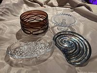 Lot of 4 serving dishes on cloth background: brown textured glass bowl, clear pedestal cake stand, Mikasa crystal candy dish, iridescent shell-shaped glass plate.