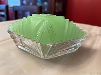 Side angle of green jadeite glass puff box lid sitting in clear glass base on tablecloth, showing stepped design and fan motifs.