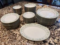 Full set of Noritake Spring Meadow dinnerware arranged on a granite countertop, showing multiple stacks of plates, bowls, and the oblong platter in front.