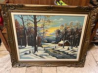 Frontal view of entire painting showing snow-covered winter landscape, flowing stream, trees, houses and sunset sky in carved wooden frame.