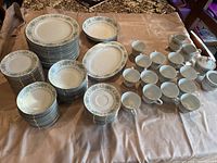 A large set of white porcelain dinnerware with a floral garland design stacked and arranged on a table. Includes plates, bowls, cups, saucers, creamers, and sugar bowls.