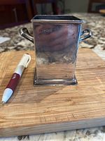 Square silver handled cup placed on wooden board with white and red pen for scale, showing tarnished surface and handles.