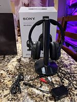 Sony WH-RF400 wireless headset displayed on black vertical base station with original box behind it. Several audio cables and a power adapter also visible on the granite countertop.