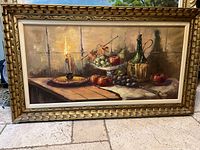 Full view of the framed oil painting showing the still life scene, candle, fruits including grapes, pear, apples, and persimmons, and a green bottle with wicker covering on a wooden table under a window.