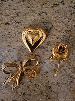 All three gold-tone brooches (heart, bow, rose) shown on granite surface.