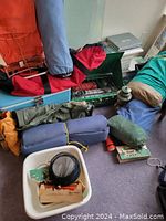 Photo of several camping items including rolled sleeping pads, the green metal gas cook stove, a white plastic utility bin with accessories, and other gear.