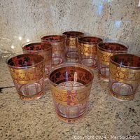 Eight clear drinking glasses arranged in a cluster showing the golden grape motif and gold decorative bands.
