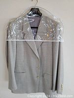 Full view of men's blazer hanging in protective garment bag, showing light grey check patterned fabric, two front flap pockets, and three-button closure.