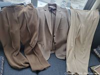 Three clothing items laid out: a dark brown pair of pants, a cream colored Armani jacket, and a light beige/cream pair of pants