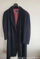 Full front view of the men's winter coat hanging on a hanger showing dark color, classic long overcoat style with lapel collar and double-breasted button closure.